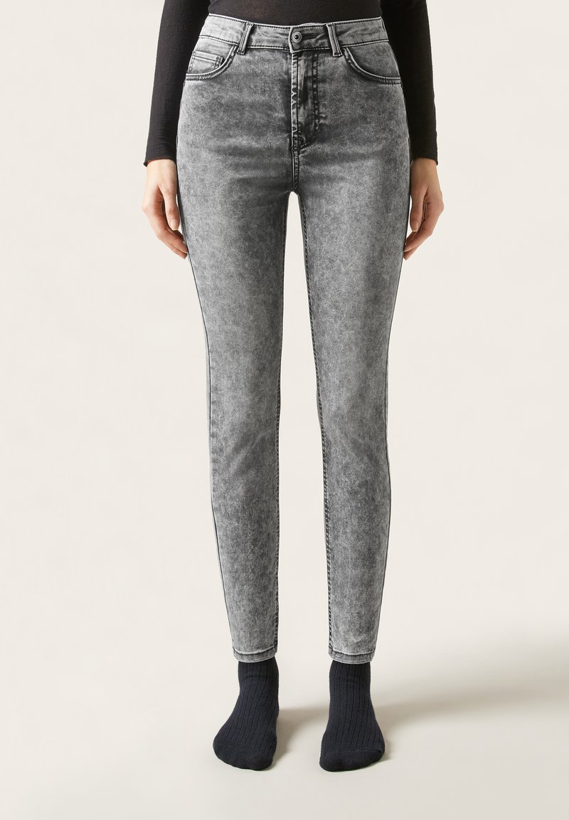 Kobieta ubrana w szare, wysokogatunkowe, obcisłe jeansy z efektem sprania oraz czarne skarpetki, stojąca na tle jednolitego tła.