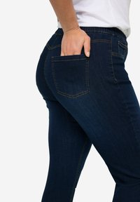 Vaqueros de mezclilla azul oscuro con un diseño ajustado, que cuentan con un bolsillo trasero, cinturilla elástica y detalles de costura sutiles.