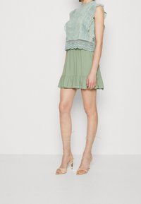 Robe verte évasée avec un corsage en dentelle, bords festonnés et ourlet en volants ; accompagnée de sandales à talons nu et à lanières.