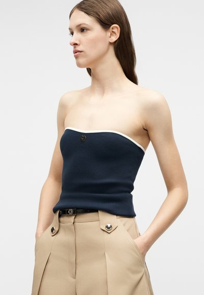 Γυναίκα που φοράει μπλε σκούρο τοπ χωρίς τιράντες με λευκό περίγραμμα και μπεζ ψηλόμεσο παντελόνι, στέκεται με τα χέρια στις τσέπες, κοιτάζοντας προς τα αριστερά.