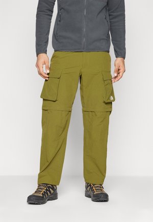 Zielone spodnie cargo wykonane z lekkiego materiału z wieloma bocznymi kieszeniami, w połączeniu z ciemnym obuwiem z pomarańczowymi akcentami.