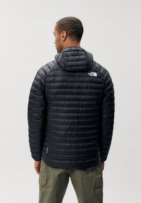Zwarte geïsoleerde pufferjack met een capuchon, met een gestikte ontwerp en logo op de bovenrug. Draag over olijfgroene cargobroek.