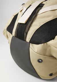 Khakifarbener Stoffrucksack mit schwarzen Mesh-Akzenten, runder Form, strapazierfähigem Lederboden und verstellbaren Trägern. Verfügt über einen Verschlussmechanismus.