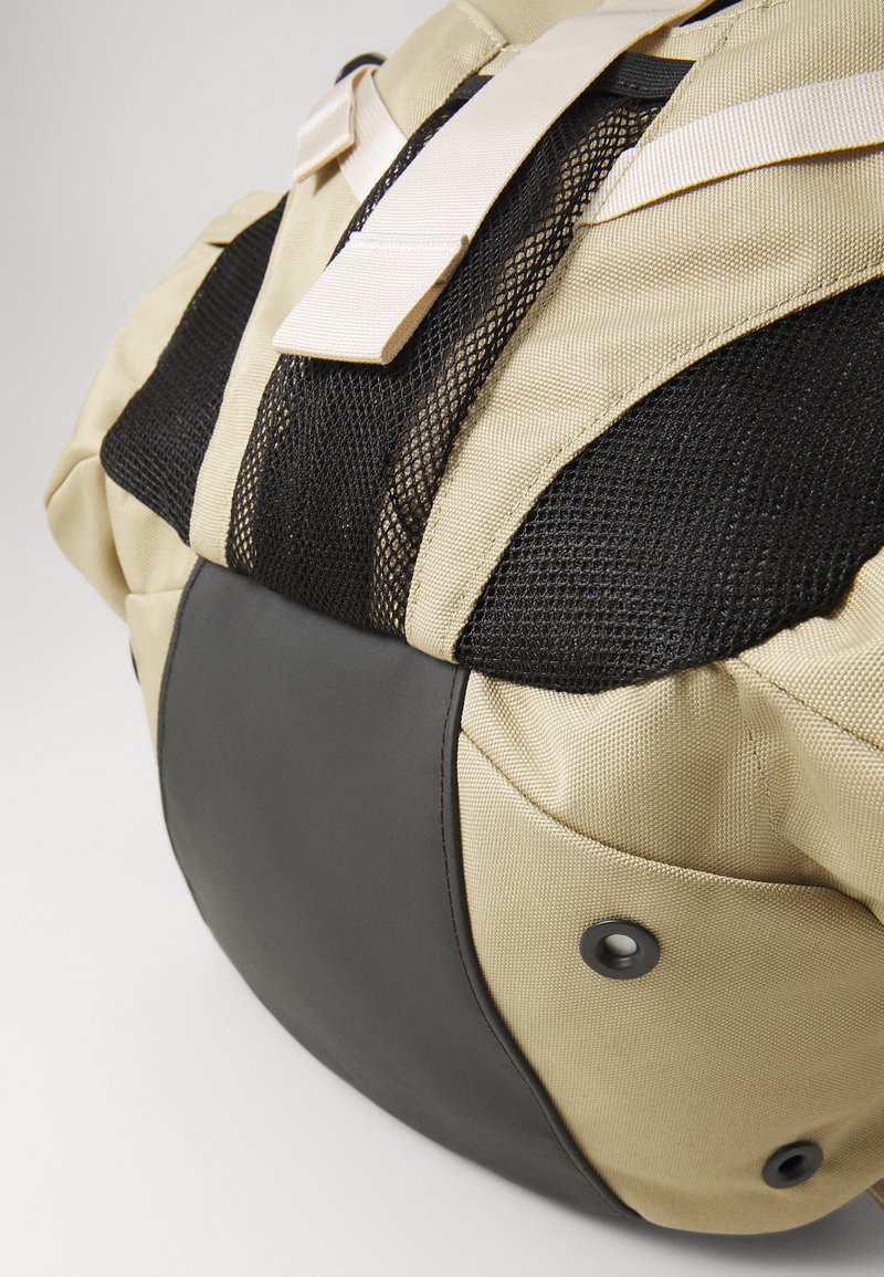 Khakifarbener Stoffrucksack mit schwarzen Mesh-Akzenten, runder Form, strapazierfähigem Lederboden und verstellbaren Trägern. Verfügt über einen Verschlussmechanismus.