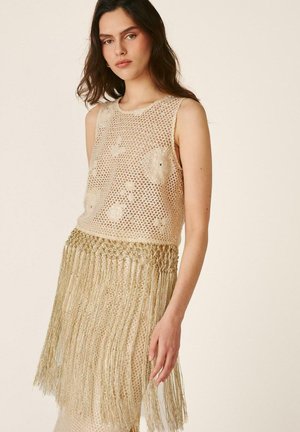 Mujer con un top de crochet beige sin mangas y falda a juego con flecos largos que cuelgan de la cintura.