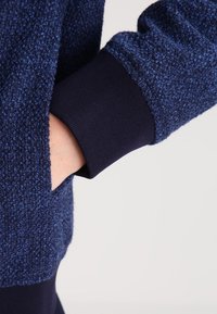 Marineblauer strukturierter Pullover mit einer samtigen Oberfläche, der einen dunklen Bund und eine Seitentasche aufweist und ein kontrastierendes glattes Gewebe zeigt.