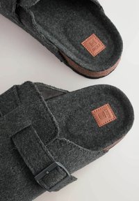 Grau gefälschte Clogs mit einem Riemenverschluss, strukturierten Oberflächen und einem braunen Lederetikett mit dem "NEXT"-Logo auf der Innensohle. Flaches, offenes Design.