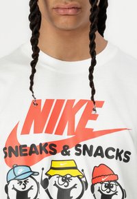Hvit bomulls T-skjorte med en stor rød Nike-logo og tegneseriefigurer iført blå, gule og røde hatter. Teksten sier "SNEAKS & SNACKS."