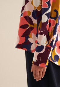 Camicia a maniche lunghe con stampa floreale colorata, caratterizzata da un vivace mix di rosa, viola e giallo. Il polsino presenta un dettaglio con bottone. Tessuto dalla consistenza morbida.