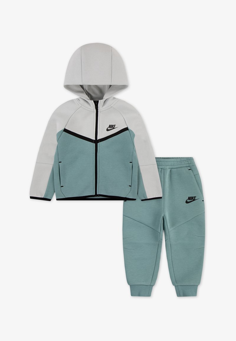 Nike treniruoklių kostiumas, turintis gobtuvą ir švarką su užtrauktuku šviesiai pilkos ir žalio mėlyno spalvų, su juodomis detalėmis, derinamas su atitinkamomis žalio mėlyno spalvos siaurėjančiomis kelnėmis.
