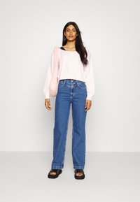 Sweatshirt cropped rosa claro com logótipo, combinado com jeans de perna larga azuis. O modelo usa sandálias pretas tipo slide e segura uma mala rosa fofa.