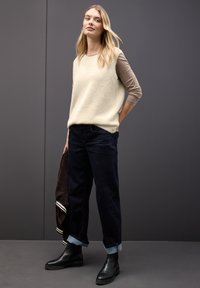 Pull en polaire crème sans manches, chemise à manches longues marron en dessous, jeans larges bleu marine avec ourlet retroussé, bottes noires jusqu'à la cheville, tenant une veste marron.