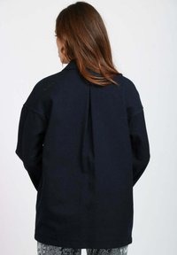 Giacca oversize blu navy con colletto, spalle scese e dettaglio di piega sul retro. Realizzata in un tessuto morbido e strutturato.