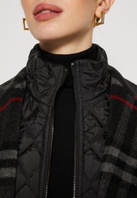 Primer plano de una persona que lleva una chaqueta negra acolchada con cremallera, un cuello alto negro y pendientes dorados geométricos.