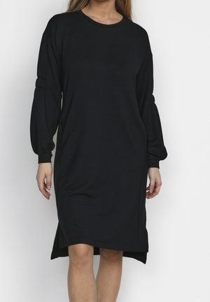 Femme portant une robe noire à manches longues, longueur genou, avec des manches froncées et un col rond, debout devant un fond clair.
