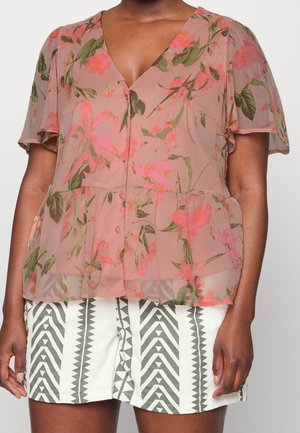 Femme portant un chemisier floral rose transparent avec des manches courtes volantées et un short blanc présentant un motif géométrique en zigzag noir.