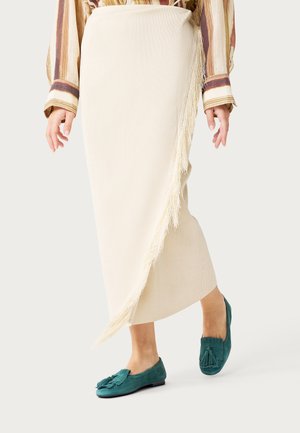 Mujer con una falda cruzada de canalé color crema con detalles de flecos y mocasines de ante color teal con borlas, posando frente a un fondo liso.