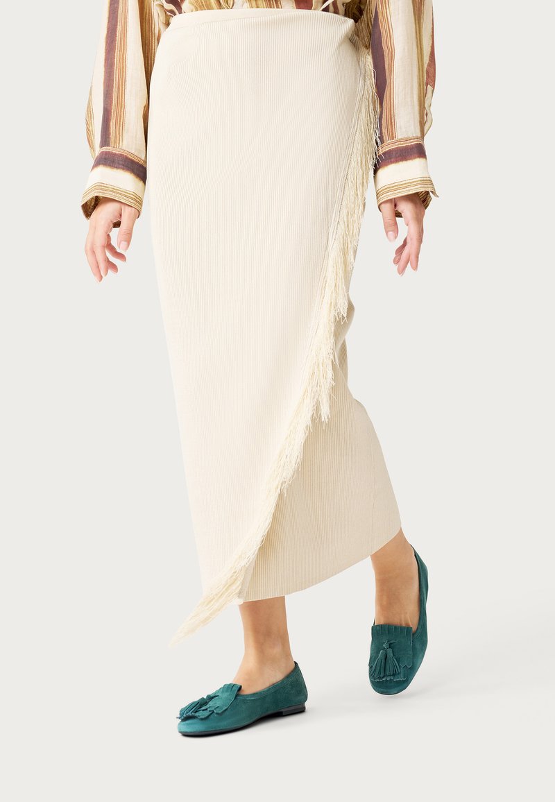Femme portant une jupe portefeuille crème côtelée avec des détails à franges et des mocassins en daim turquoise à glands, debout devant un fond uni.