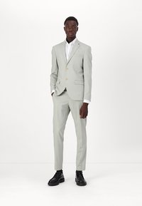 Šviesiai pilkas kostiumas su dviejų mygtukų švarku ir tiesiais keliais. Kartu su baltu marškinėliu ir juodais šnaruojamais batais, turintis lygų paviršių.