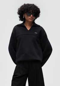 Felpa nera oversize con scollo a V e colletto con mezza zip, con una cucitura orizzontale e un piccolo logo con patch. Tessuto morbido.