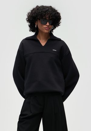 Felpa nera oversize con scollo a V e colletto con mezza zip, con una cucitura orizzontale e un piccolo logo con patch. Tessuto morbido.