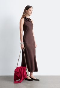 Braunes ärmelloses Kleid mit hohem Kragen, glatter Textur und figurbetontem Schnitt. Die Accessoires bestehen aus einem roten Schal und einer hellen Tasche. Schwarze Ballerinas.