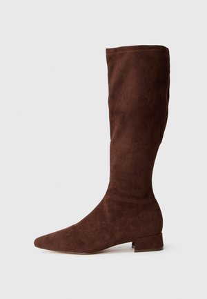 Botas de ante marrón hasta la rodilla con punta alargada y un tacón bajo tipo bloque. Los detalles en la costura lateral añaden una textura sutil.