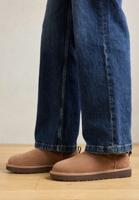 Ruskeanan mokkanahkasaappaat, joissa on tasapohja, pyöreä kärki ja pieni vetonauha, yhdistettyinä sinisiin denim-farkkuihin, joissa on suoralinjainen leikkaus.