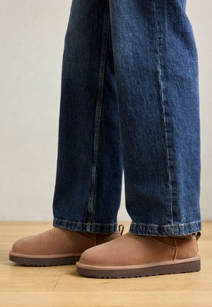 Bottines en daim marron avec une semelle plate, un bout arrondi et une petite languette, associées à un jean en denim bleu de coupe droite.