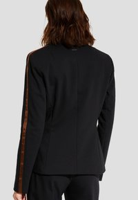 Blazer negro con mangas largas, con un ajuste slim y detalle de franjas laterales de terciopelo marrón a lo largo de las mangas y la espalda. Textura suave, sin forro.