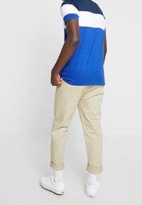 Muž oblečený v modro-bílé polo triko s barevně oddělenými částmi, béžových kalhotách s ohrnutými nohavicemi, bílých ponožkách a bílých teniskách, stojící s zády otočenými dopředu.