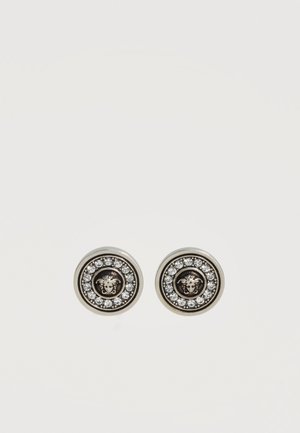 Boucles d'oreilles rondes en argent avec un emblème central de tête de Méduse, entouré d'un cercle de petites pierres précieuses claires sur fond blanc.