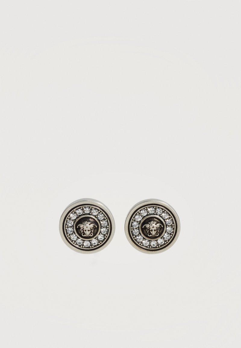 Boucles d'oreilles rondes en argent avec un emblème central de tête de Méduse, entouré d'un cercle de petites pierres précieuses claires sur fond blanc.