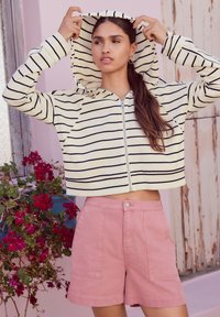 Afgeknipte hoodie in crème met zwarte strepen, ritssluiting en capuchon. Gecombineerd met roze korte broek met voorzakken.