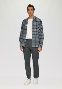 Man in een zwart-wit geblokt overhemd over een wit T-shirt, grijze broek en witte sneakers, staand tegen een lichte achtergrond.