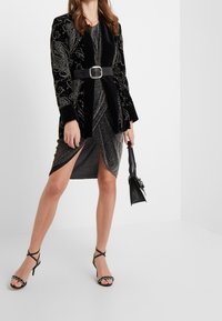 Czarna aksamitna marynarka z srebrnym haftem w kwiaty, zestawiona z wzorzystą czarną i srebrną sukienką kopertową oraz czarnymi sandałami na obcasie.