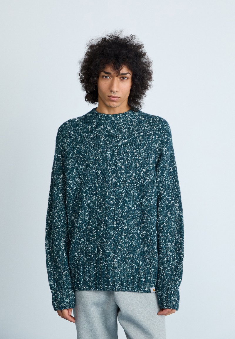 Gebreide trui in donkerteal met een textuurpatroon en een ribbelkraag. Heeft lange mouwen en een rechte zoom, gedragen over grijze broek.