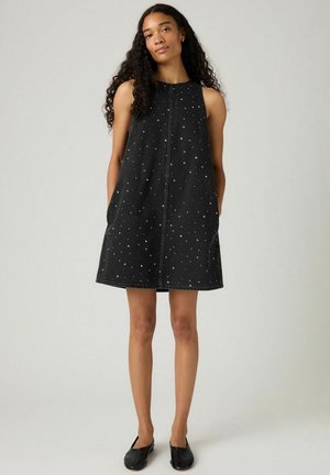 Model wearing a sleeveless black dress with silver dots, standing against a neutral background, hands in pockets, showcasing a relaxed pose.