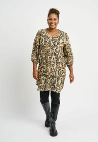 Tunika s leopard uzorkom u bež boji, sa opuštenim krojem, V-izrezom i rukavima do lakta, u kombinaciji s crnim tajicama i čizmama do koljena.