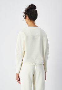 Sudadera de color crema claro con mangas largas, un corte relajado y un detalle de logo minimalista en la parte trasera. Textura suave.