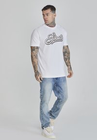 Balta T-kreklu ar melno "Siksilk" logo, apvienota ar gaiši ziliem džinsiem. Modelis valkā baltas kedas, demonstrējot ikdienišķu ielas apģērbu stilu.