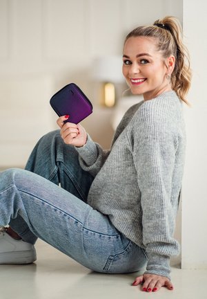 Purple-ombre zippered wallet held by a person, set against a light interior. Soft, textured fabric with a rounded shape and minimalist logo.