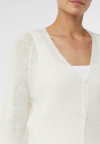 White knitted cardigan with a v-neck and long sleeves. Features a soft texture, button closure, and sheer knit detailing on the shoulders.