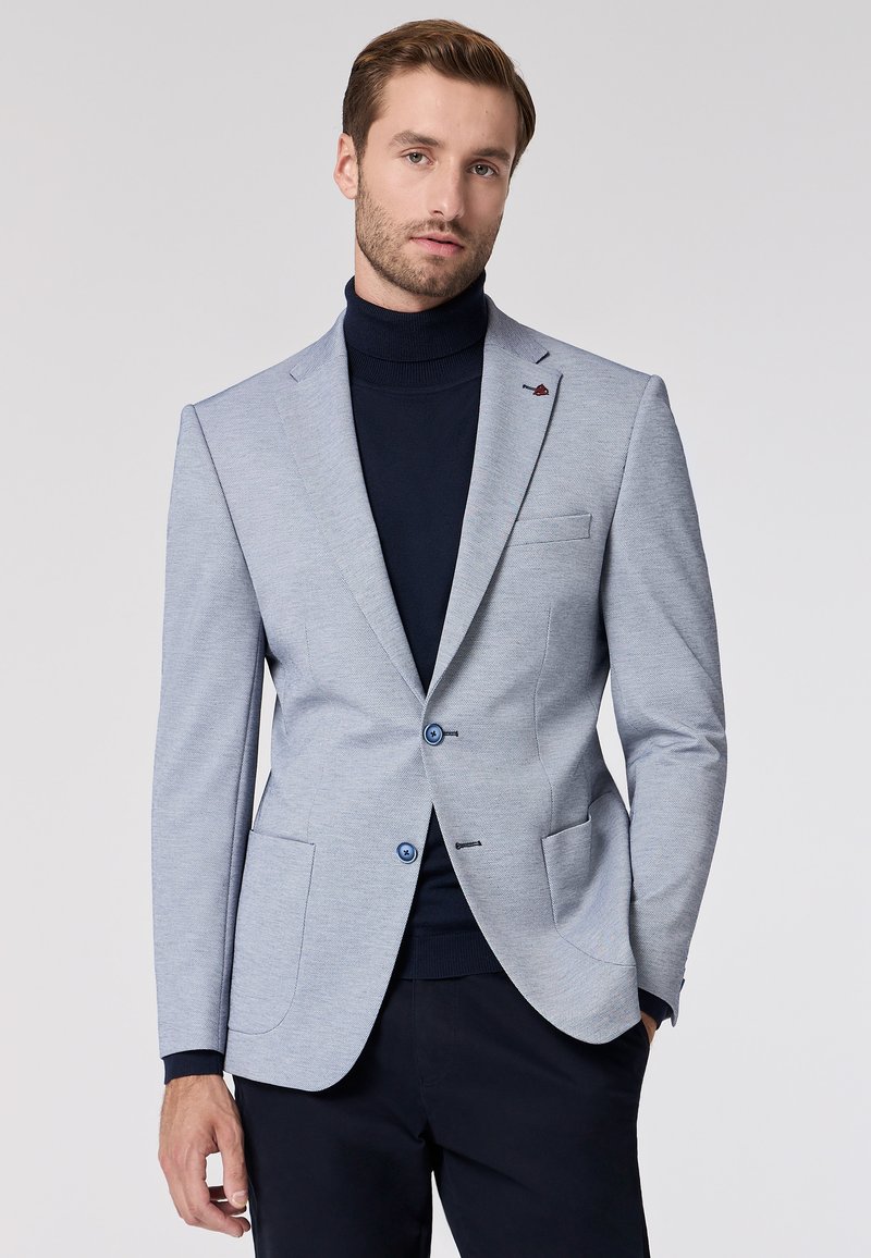 Blazer grigio chiaro con un motivo testurizzato, due bottoni blu e rever a taglio. Indossato sopra un dolcevita blu navy, dotato di due tasche frontali.
