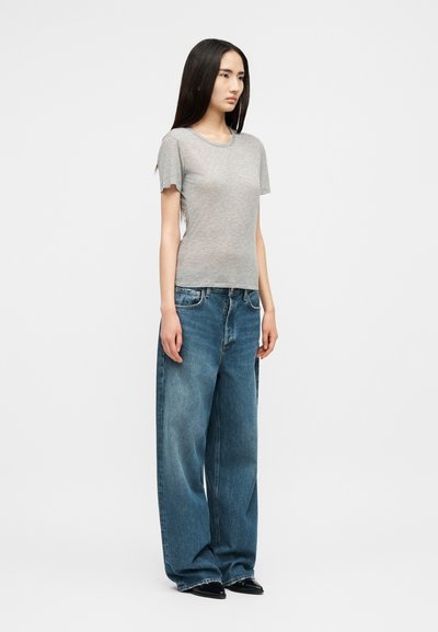 Mlada žena stoji noseći prianjajuću sivu majicu s kratkim rukavima, široke plave traperice i crne cipele špicastog vrha na jednostavnoj pozadini.