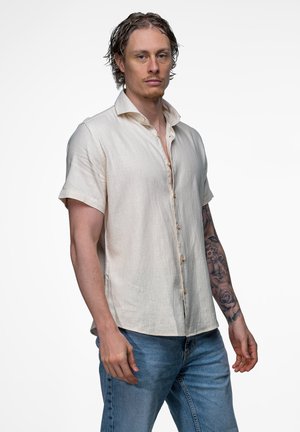 Man met krullend haar, licht kort mouw knoopshirt, blauwe spijkerbroek en een gedetailleerde tatoeage-mouw op zijn linkerarm staat naar voren gericht.