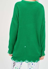 Personne portant un pull vert tricoté surdimensionné avec des détails usés, assorti à un pantalon blanc avec des éclaboussures de peinture bleues et vertes.