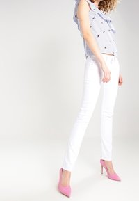 Weiße, figurbetonte Jeans kombiniert mit einem blau-weißen gestreiften Oberteil mit Rüschen und floralen Stickereien. Rosa Stilettos vervollständigen das Outfit.