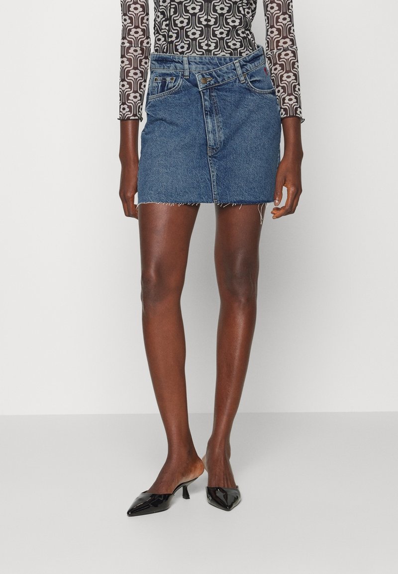 Femme portant une mini-jupe en jean asymétrique, des escarpins noirs vernis à bout pointu et un haut à manches longues à motifs, sur fond uni.