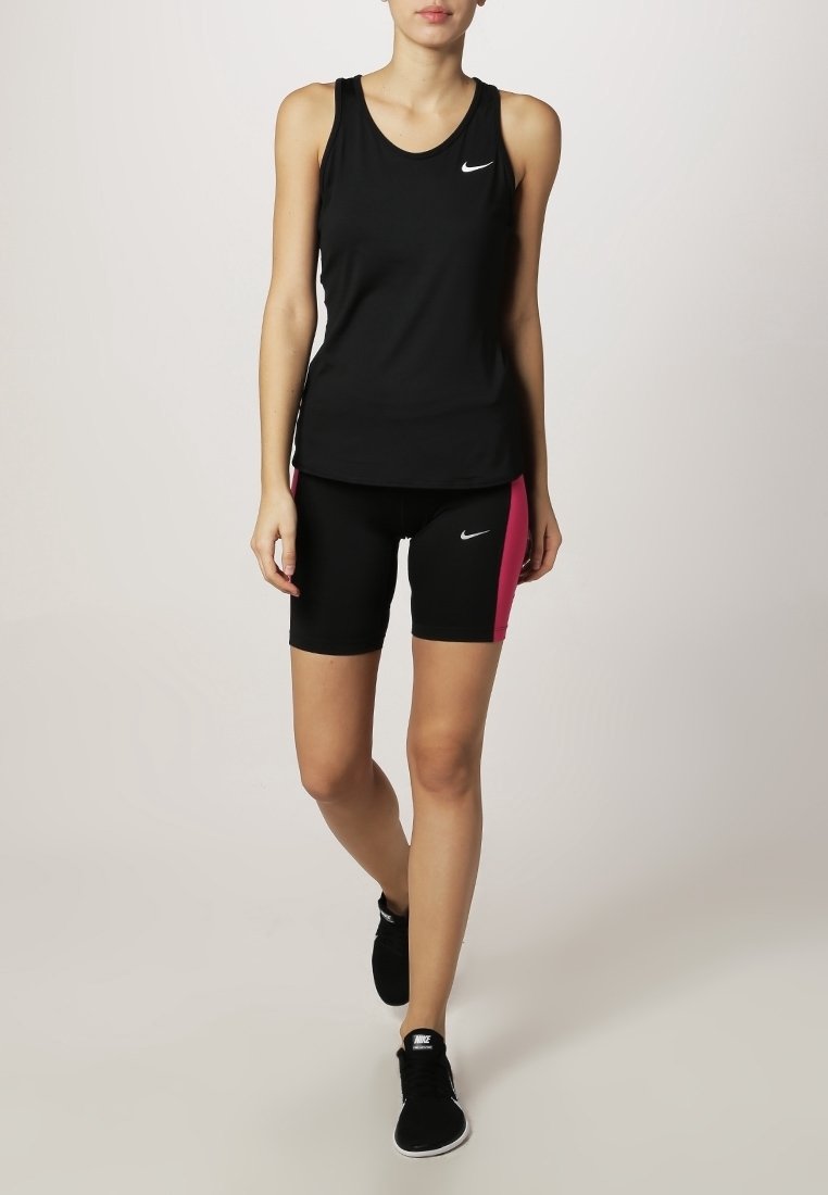 Femme marchant portant un débardeur de sport noir Nike, un short noir et rose et des baskets noires sur fond clair uni.